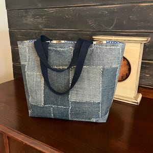 May include: A denim tote bag with a patchwork design. The bag features a rectangular shape with a dark blue handle and a patterned interior lining. The denim is a mix of light and medium blue shades.