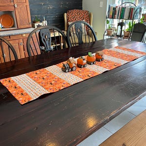 May include: A dark wooden dining table is set with a Halloween-themed table runner and decorative pumpkin train figurines. The runner features orange and white stripes with Halloween patterns. The table is surrounded by black chairs.