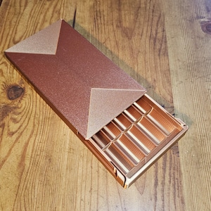 May include: A copper-coloured rectangular box with a lid featuring a triangular design. The box is open, revealing a series of rectangular compartments. The box rests on a wooden surface.