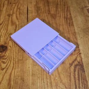May include: A lavender-colored, rectangular organizer with a sliding lid. The interior is divided into several compartments of varying sizes. The organizer is open, revealing the interior compartments, and sits on a wooden surface.