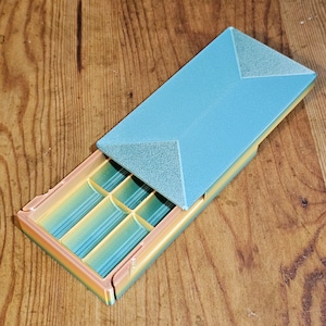 May include: A 3D-printed box with a light blue lid and a multi-colored base. The base has several compartments, and the lid is slightly ajar. The box is made of plastic and sits on a wooden surface.