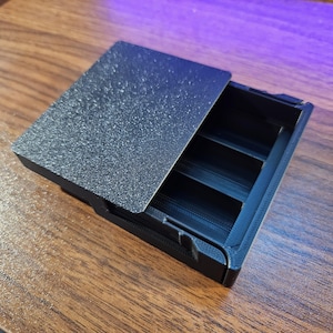 May include: A black, rectangular storage box with a textured lid, partially open to reveal three interior compartments. The box is made of a matte material and sits on a wooden surface.