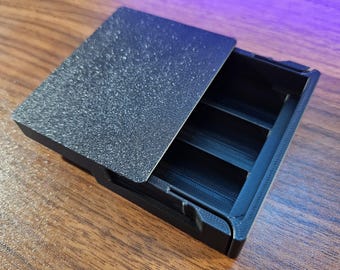 May include: A black, rectangular storage box with a textured lid, partially open to reveal three interior compartments. The box is made of a matte material and sits on a wooden surface.