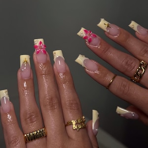 May include: A set of long, square-shaped artificial nails. The nails feature a light yellow tip with a pink and clear base. Some nails are adorned with pink flower designs, gold stars, and small pearls. Gold rings are worn on the fingers.