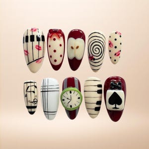 Puede incluir: Una colección de diez diseños de uñas en forma de almendra. Los diseños incluyen un teclado de piano, lunares, una manzana, una espiral, notas musicales, un reloj y un símbolo de carta de juego. Las uñas están pintadas en varios colores.