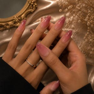 Puede incluir: Manos con uñas largas y ovaladas con un diseño degradado, que transiciona de una base transparente a un tono rosa intenso. Un anillo de oro con pequeñas piedras transparentes adorna un dedo. Las uñas están bien cuidadas y pulidas.