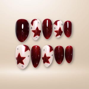 May include: A set of ten artificial nails in a variety of designs. Some nails are solid burgundy, while others feature a white base with a burgundy star and small rhinestones. The nails are oval-shaped.