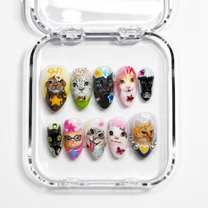 May include: A collection of ten artificial nails, each decorated with a unique cat face design. The nails are presented in a clear plastic case. The cat faces feature diverse colours and styles, including a black cat, a tabby, and a calico cat.