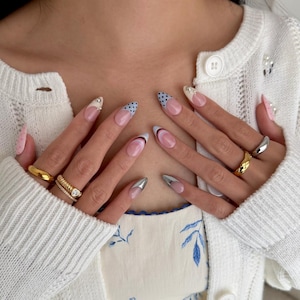 May include: A close-up of manicured nails with various designs. Some nails feature a silver metallic finish, while others have a French tip with blue polka dots or a red and black curved design. Gold rings adorn the fingers.