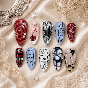 Könnte beinhalten: Eine Sammlung von zehn künstlichen Nägeln mit verschiedenen Designs. Die Nägel weisen Farben wie Rot, Schwarz, Blau und Nude auf, mit Verzierungen wie Strasssteinen und sternförmigen Akzenten. Designs umfassen Wirbel, Kirschen, Sterne und Cartoon-Gesichter.