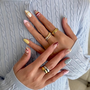 May include: Close-up of hands displaying diverse nail art designs. The nails showcase patterns including polka dots, stripes, and plaid in colours like yellow, red, pink, and blue. Gold and silver rings are worn on the fingers. The background is a light blue cable knit jumper.