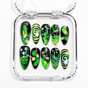 May include: A set of ten press-on nails with a clear case. Each nail features a unique design with black, green, and silver accents. Designs include alien and cat faces, swirls, and stars. The nails are oval-shaped.