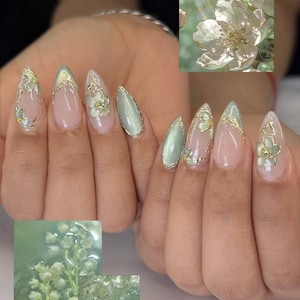 May include: Almond-shaped nails with a professional manicure. The nails have a light pink base, gold trim, and floral designs. Some nails are a soft green. The overall look is elegant and feminine, with a touch of nature-inspired artistry.