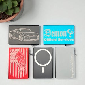 May include: A collection of five metal cardholders with various designs. One features a car outline, another has "Demon Oilfield Services" in turquoise, and a third displays an American flag and eagle. The remaining two have a circle and the text "I love you forever and always".