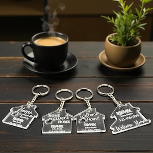 May include: Four clear acrylic house-shaped keychains with silver keyrings. Each keychain has a different message: "Home is where the heart is", "Home sweet Home", "Welcome Home!", and "Welcome Home". Each keychain has the RE/MAX logo and a phone number.