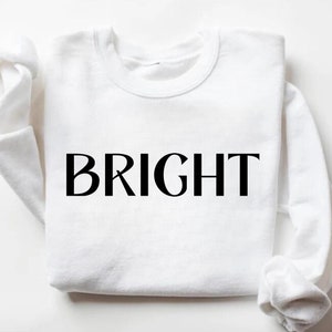May include: A white crew neck sweatshirt with the word "BRIGHT" printed in large, black capital letters across the front. The sleeves are folded up. The sweatshirt is made of a soft material.