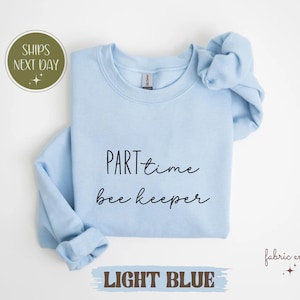 May include: Light blue crewneck sweatshirt with the text "PART time bee keeper" in black script. The sweatshirt has rolled sleeves and the words "LIGHT BLUE" are printed at the bottom.