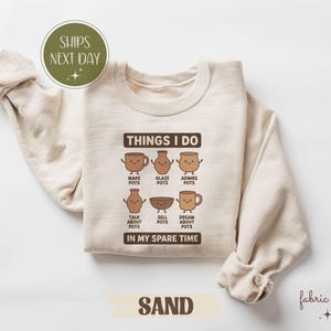 May include: A sand-coloured sweatshirt featuring the text "THINGS I DO" and pottery illustrations. The text "IN MY SPARE TIME" is below. The sweatshirt also has the words "SHIPS NEXT DAY" in a green circle.