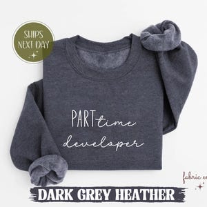 Op de afbeelding: Donkergrijze heather sweatshirt met de tekst "part time developer" in witte letters. De sweatshirt heeft een ronde hals en lange mouwen. De woorden "Dark Grey Heather" staan onderaan de afbeelding.