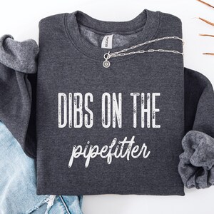 以下が含まれることがあります： 「DIBS ON THE pipefitter」の白文字がプリントされた濃いグレーのスウェットシャツ。ペンダント付きのシルバーネックレスがスウェットシャツの上にかけられています。袖は折り返されており、明るいグレーの裏地が見えています。ジーンズが見えています。