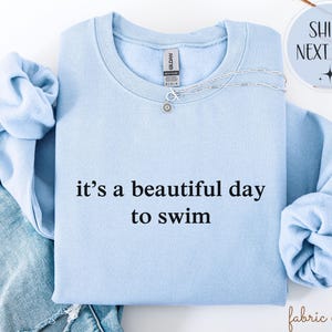 May include: A pale blue sweatshirt featuring the phrase "it's a beautiful day to swim" in black text. A silver necklace with a sun pendant is on the sweatshirt. The sleeves are tucked in.