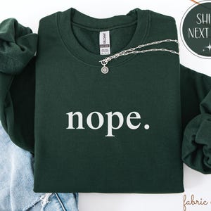 May include: A dark green sweatshirt with the word "nope" in white, paired with a silver necklace. The sweatshirt is folded on top of a pair of blue jeans. The image also includes a "Ships Next Day" graphic.