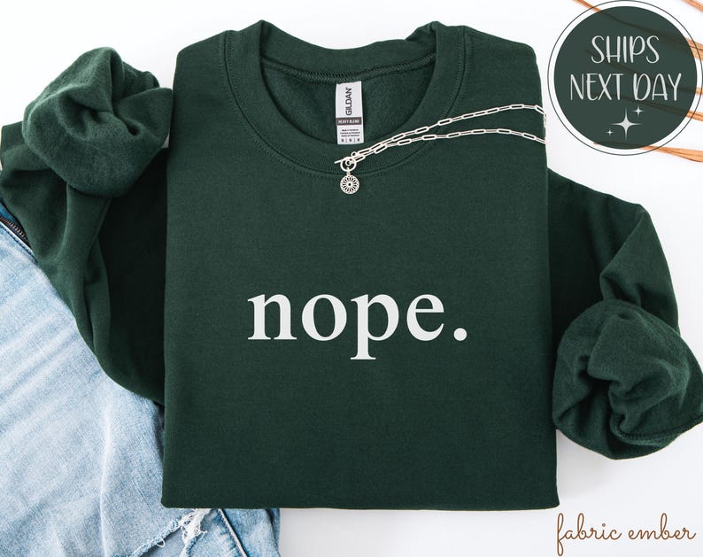 May include: A dark green sweatshirt with the word "nope" in white, paired with a silver necklace. The sweatshirt is folded on top of a pair of blue jeans. The image also includes a "Ships Next Day" graphic.