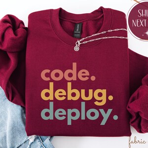 May include: A maroon sweatshirt featuring the text "code. debug. deploy." in a stacked, multi-coloured font. A silver necklace with a small pendant is placed on the sweatshirt. The image also includes a "Ships Next Day" graphic.
