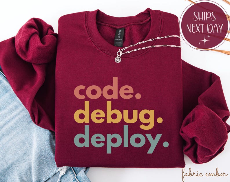 May include: A maroon sweatshirt featuring the text "code. debug. deploy." in a stacked, multi-coloured font. A silver necklace with a small pendant is placed on the sweatshirt. The image also includes a "Ships Next Day" graphic.