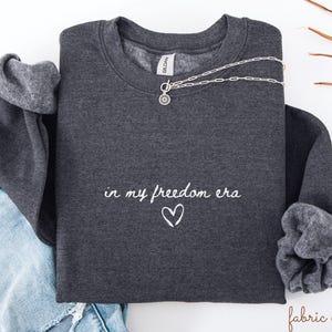 Op de afbeelding: Een donkergrijs sweatshirt met de tekst "in my freedom era" en een hartje in witte letters. Een zilveren ketting met een hanger is over de voorkant gedrapeerd. Het sweatshirt is op een spijkerbroek gevouwen.