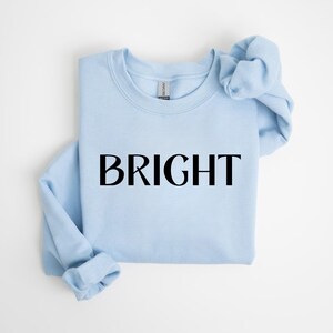 May include: A pale blue sweatshirt featuring the word "BRIGHT" in black lettering. The garment is neatly folded, with the sleeves and neckline visible. The background is a plain white surface.