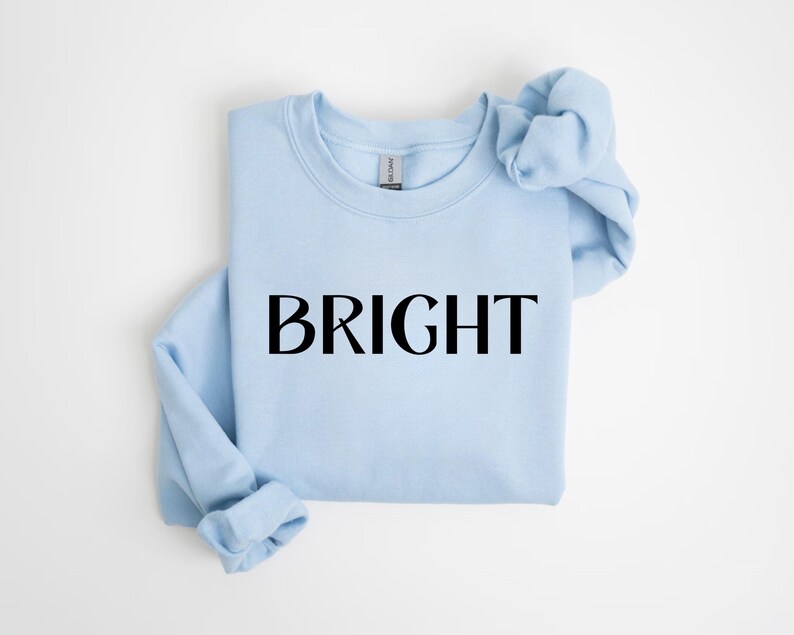 May include: A pale blue sweatshirt featuring the word "BRIGHT" in black lettering. The garment is neatly folded, with the sleeves and neckline visible. The background is a plain white surface.