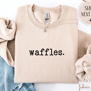 K&ouml;nnte beinhalten: Beiges Sweatshirt mit dem Wort "waffles." in Schwarz gedruckt. Eine silberne Halskette mit einem kleinen Anh&auml;nger ist &uuml;ber die Vorderseite drapiert. Das Sweatshirt ist gefaltet, die &Auml;rmel sind eingesteckt. Die Worte "Ships Next Day" stehen in einem Kreis oben rechts.