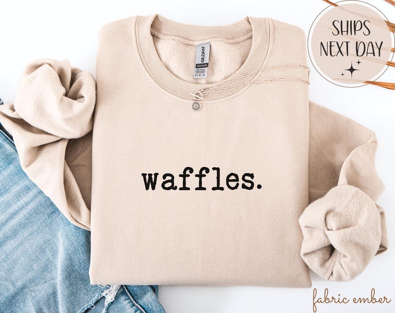 K&ouml;nnte beinhalten: Beiges Sweatshirt mit dem Wort "waffles." in Schwarz gedruckt. Eine silberne Halskette mit einem kleinen Anh&auml;nger ist &uuml;ber die Vorderseite drapiert. Das Sweatshirt ist gefaltet, die &Auml;rmel sind eingesteckt. Die Worte "Ships Next Day" stehen in einem Kreis oben rechts.