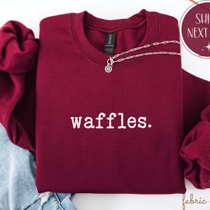 K&ouml;nnte beinhalten: Ein weinrotes Sweatshirt mit dem Wort "waffles" in Wei&szlig;, einer silbernen Halskette und einer Grafik "Versand am n&auml;chsten Tag". Das Sweatshirt ist auf einer blauen Jeans gefaltet. Die &Auml;rmel sind zur Seite gefaltet.