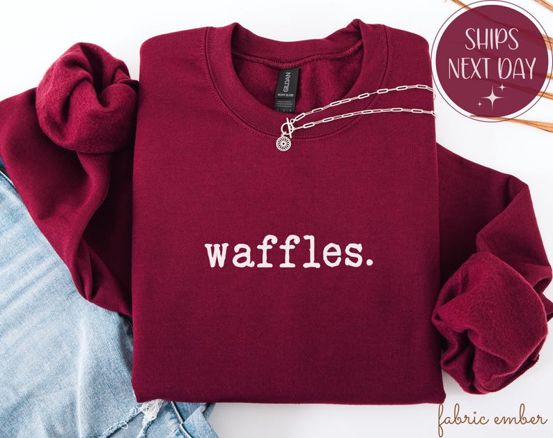 K&ouml;nnte beinhalten: Ein weinrotes Sweatshirt mit dem Wort "waffles" in Wei&szlig;, einer silbernen Halskette und einer Grafik "Versand am n&auml;chsten Tag". Das Sweatshirt ist auf einer blauen Jeans gefaltet. Die &Auml;rmel sind zur Seite gefaltet.