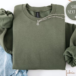 May include: An olive green sweatshirt with a crew neck and long sleeves. A silver necklace with a circular pendant is draped across the front. The sweatshirt is folded on top of a pair of blue jeans.