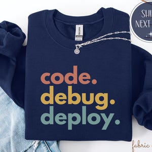 May include: A navy blue sweatshirt featuring the text "code. debug. deploy." in orange, yellow, and teal. A silver necklace with a pendant is placed on the folded sweatshirt. The sweatshirt is on top of blue jeans.