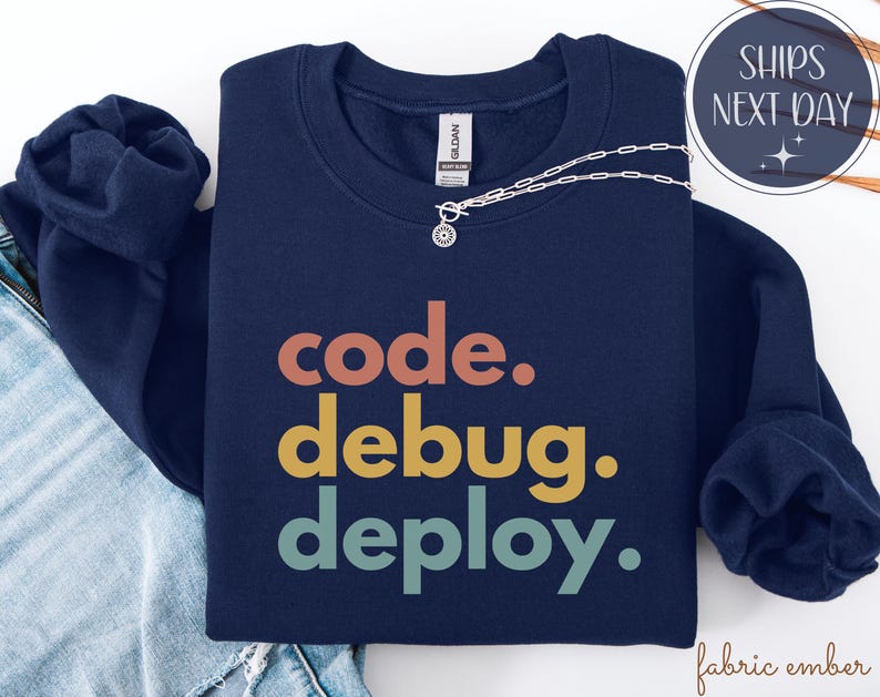 May include: A navy blue sweatshirt featuring the text "code. debug. deploy." in orange, yellow, and teal. A silver necklace with a pendant is placed on the folded sweatshirt. The sweatshirt is on top of blue jeans.