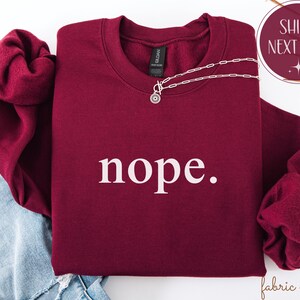 May include: A burgundy crewneck sweatshirt with the word "nope" printed in white. A silver necklace with a circular pendant is draped across the front. The sweatshirt is folded on top of blue denim.