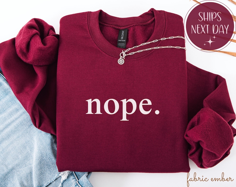 May include: A burgundy crewneck sweatshirt with the word "nope" printed in white. A silver necklace with a circular pendant is draped across the front. The sweatshirt is folded on top of blue denim.