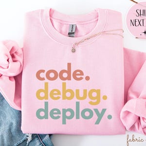 May include: A pale pink sweatshirt featuring the words "code. debug. deploy." in brown, yellow, and teal lettering. A gold necklace with a small pendant is placed on the sweatshirt. The sweatshirt is folded on top of a pair of blue jeans.