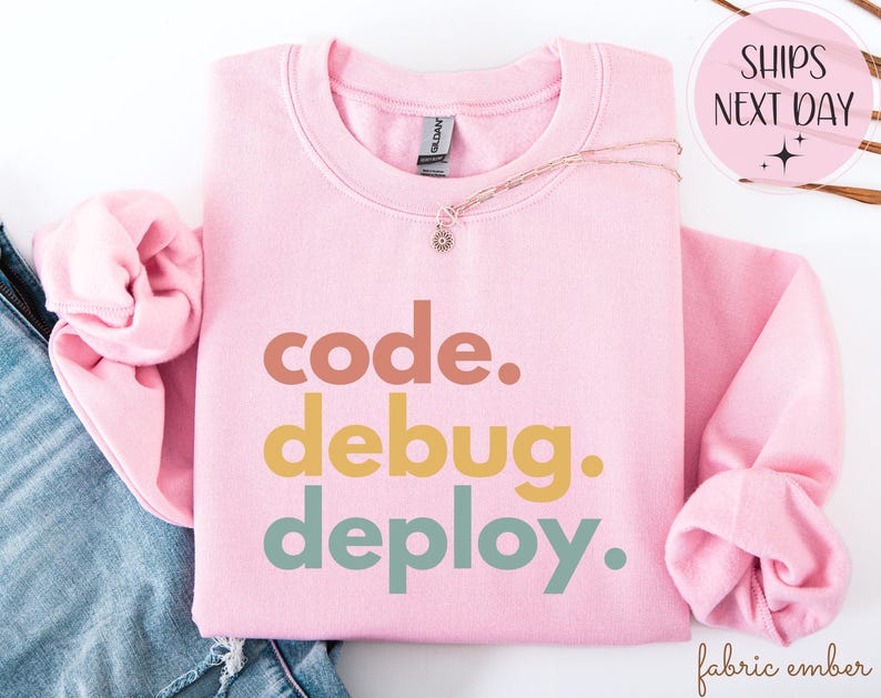 May include: A pale pink sweatshirt featuring the words "code. debug. deploy." in brown, yellow, and teal lettering. A gold necklace with a small pendant is placed on the sweatshirt. The sweatshirt is folded on top of a pair of blue jeans.