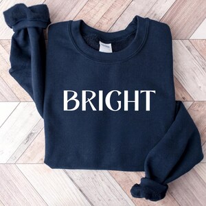 May include: A navy blue sweatshirt with the word "BRIGHT" printed in large, white capital letters across the front. The sweatshirt has a crew neck and long sleeves with cuffed wrists. The garment is made of a soft material.