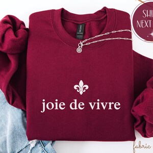 May include: A maroon crewneck sweatshirt with the phrase "joie de vivre" in white, and a fleur-de-lis symbol. A silver necklace with a pendant is draped across the sweatshirt. The sweatshirt is folded on top of a pair of blue jeans.