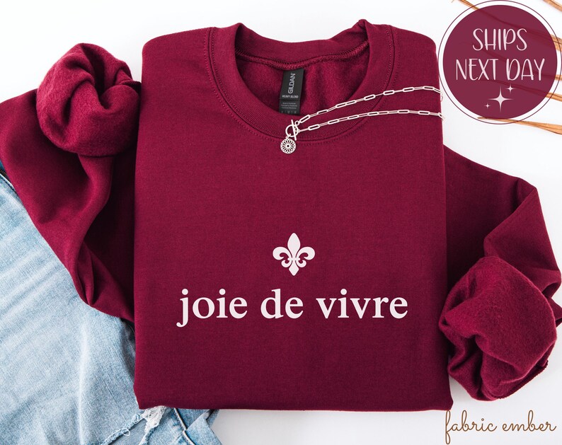 May include: A maroon crewneck sweatshirt with the phrase "joie de vivre" in white, and a fleur-de-lis symbol. A silver necklace with a pendant is draped across the sweatshirt. The sweatshirt is folded on top of a pair of blue jeans.