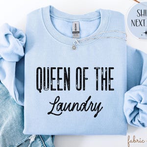 May include: A light blue sweatshirt with the text "QUEEN OF THE LAUNDRY" in black. A silver necklace with a small pendant is on the sweatshirt. The sweatshirt is folded on top of a pair of blue jeans.