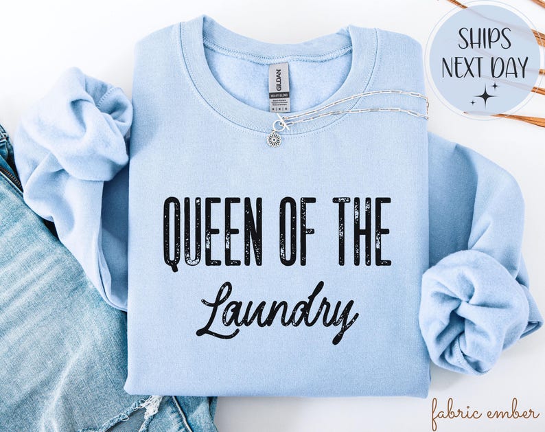 May include: A light blue sweatshirt with the text "QUEEN OF THE LAUNDRY" in black. A silver necklace with a small pendant is on the sweatshirt. The sweatshirt is folded on top of a pair of blue jeans.