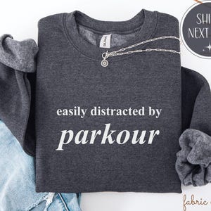 May include: A dark gray sweatshirt with the text "easily distracted by parkour" in white. A silver necklace with a pendant is draped across the shirt. The sleeves are folded, and a pair of blue jeans are visible.