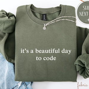 May include: An olive green sweatshirt with the white text "it's a beautiful day to code". A silver necklace with a circular pendant is draped across the front. The sweatshirt is folded, with the sleeves visible.